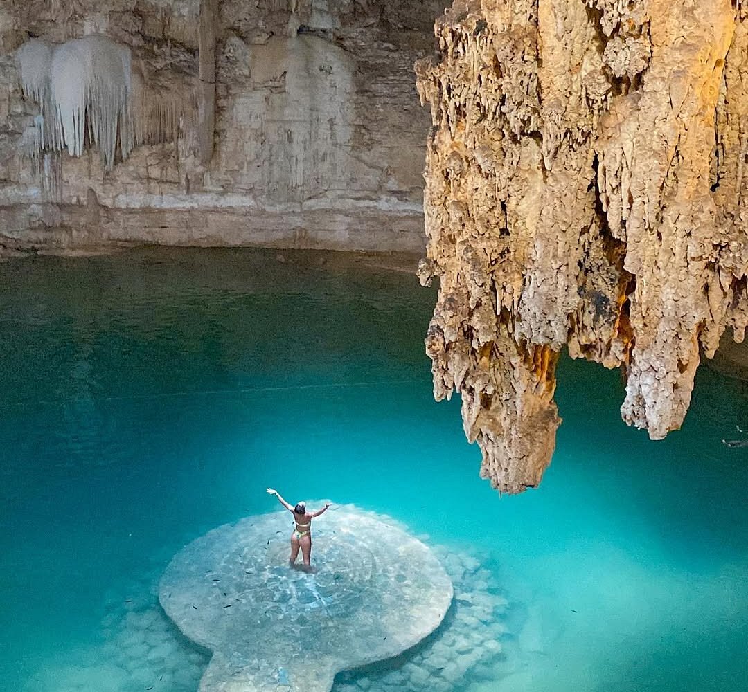 A visitor on the Suytun platform beneath a mass of stalactites, turquoise water around her