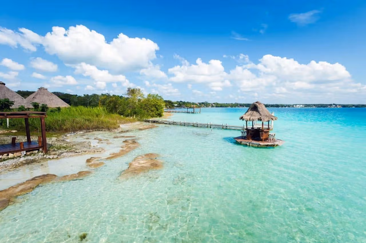 A thatched palapa on a wooden pier extending over the clear, multi-toned water of the Bacalar lagoon