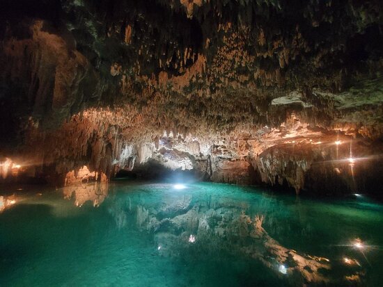 The interior of Cenote El Sueño, stalactites descending from the cavern roof above still green water lit from within