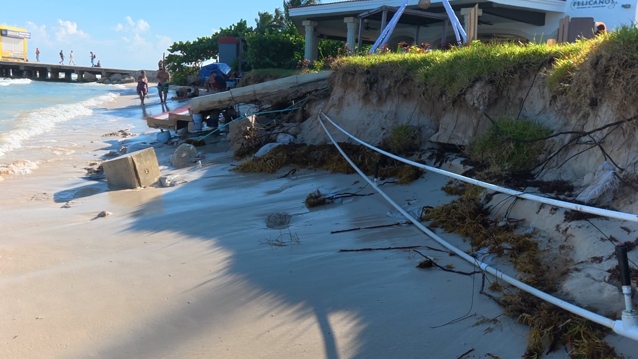 A collapsed concrete stairway and exposed pipes on the eroded beach at Playa Constituyentes, with the pier behind