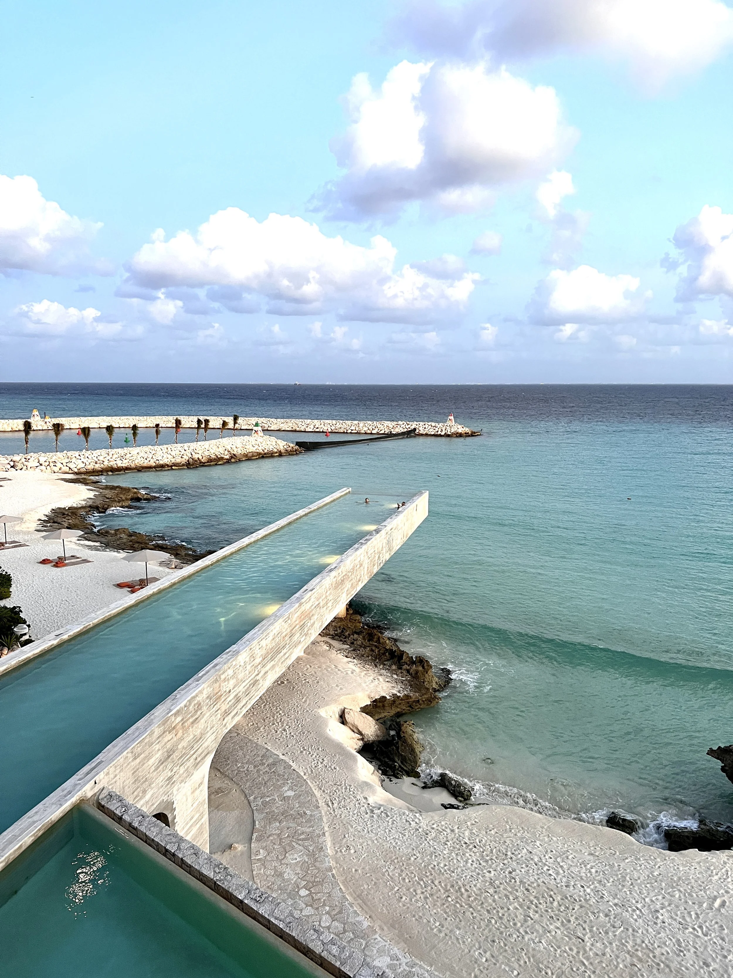 A long channel pool at golden hour extending toward a stone breakwater and open sea at La Casa de la Playa