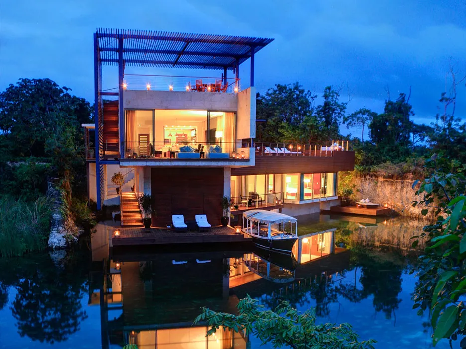A glowing modern lagoon suite reflected in the water at dusk, with a private boat moored at its dock, Rosewood Mayakoba