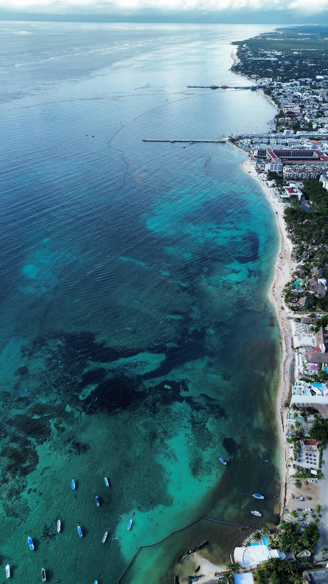 Aerial view north along the centre of Playa del Carmen on September 25, 2026, between Parque Fundadores and Calle 44, showing blue and turquoise water, the ferry piers and the containment barrier offshore
