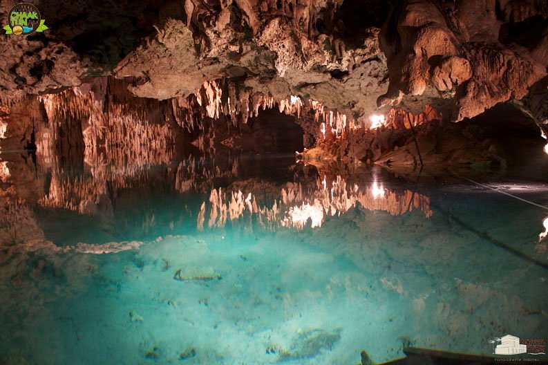 Stalactites hanging over lit turquoise water inside the cave cenote at Chaak Tun, reflected in the still surface