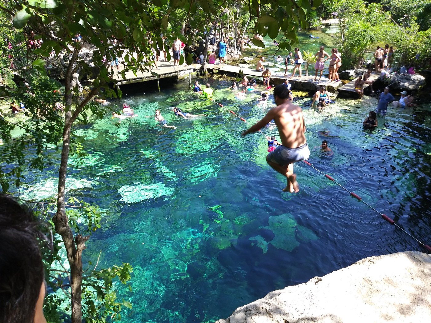 A swimmer leaping into the clear water of Cenote Cristalino, with concrete walkways and a crowd around the pool