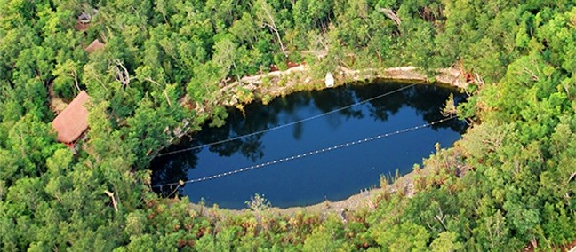 Aerial view of the dark oval pool of Cenote Las Mojarras enclosed by dense jungle, with zipline cables strung across it and palapa roofs at one end